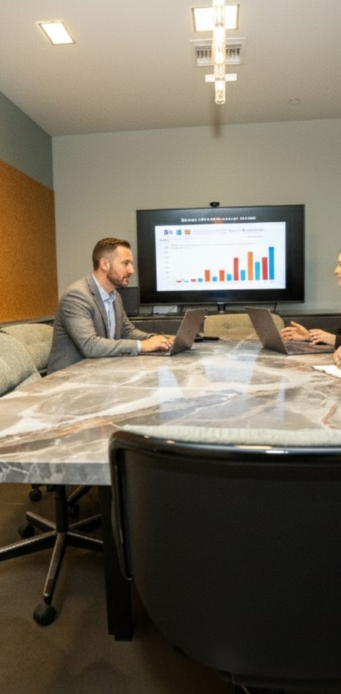 Barrister Suites Office Lease Three people in business attire sit around a conference table, looking at a laptop and a wall-mounted screen displaying a colorful bar graph during a meeting in a modern office.